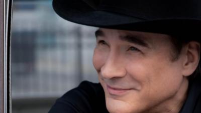 Clint Black smiles off camera as he leans out a car door. He wears a black shirt and black cowboy hat. 