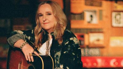 A woman with blond hair sits on a stool with her acoustic guitar on her lap, the background looks like she is in a honky tonk