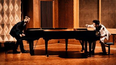 Two pianists, one dressed in white and one in black, face each other while playing a piano.
