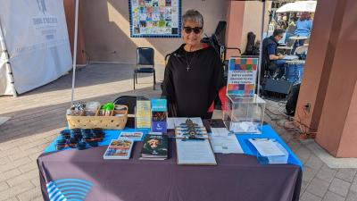 Woman in a black shirt and sunglasses standing behind a table with papers on it and a table cloth reading Chandler Center