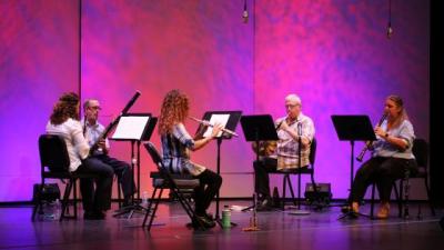 A wind quintet performs in front of a pink and purple backdrop.