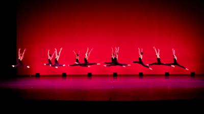 A row of ballet dancers wearing black costumes leap in front of a red background.