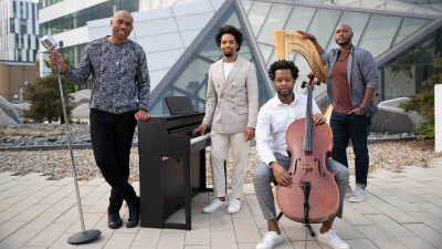 Four artists stand with their string instruments in front of a modern building