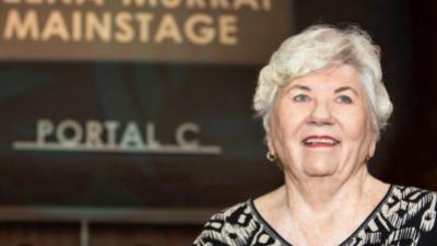 A woman with silver hair and blue eyes smiles in front of Portal C at the theatre