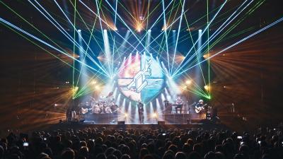 Stage with a lot of light lines shooting upward. A band on the stage and a crowd of people in front of them