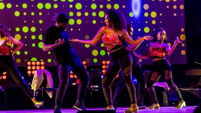 3 women and one man tap dancing, looking at each other. The lights are a purple/green and they are all wearing black and red