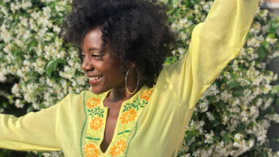 Niki J Crawford, wearing a bright yellow shirt, dances in front of a wall of white flowers.