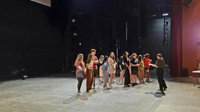 A group of students stands on a stage while a woman talks to them