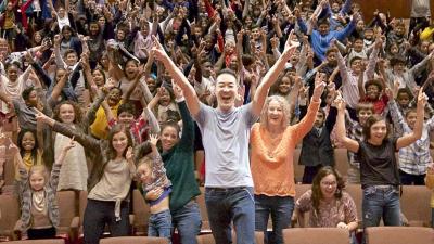 Artist Alpin Hong stands with his hands raised in joy along with a theatre full of children