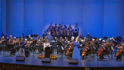 A full orchestra of musicians, conductor and two singers perform on the stage,