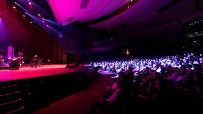 A darkened theatre, an audience watches musicians play on stage with spotlights overhead