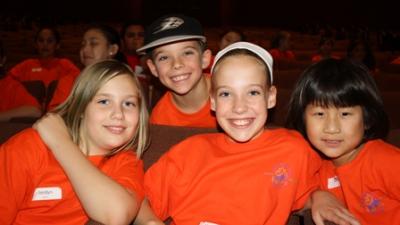 four children sit in theatre seats, smiling at the camera. All of them swear orange shirts. There are three girls and one boy. 