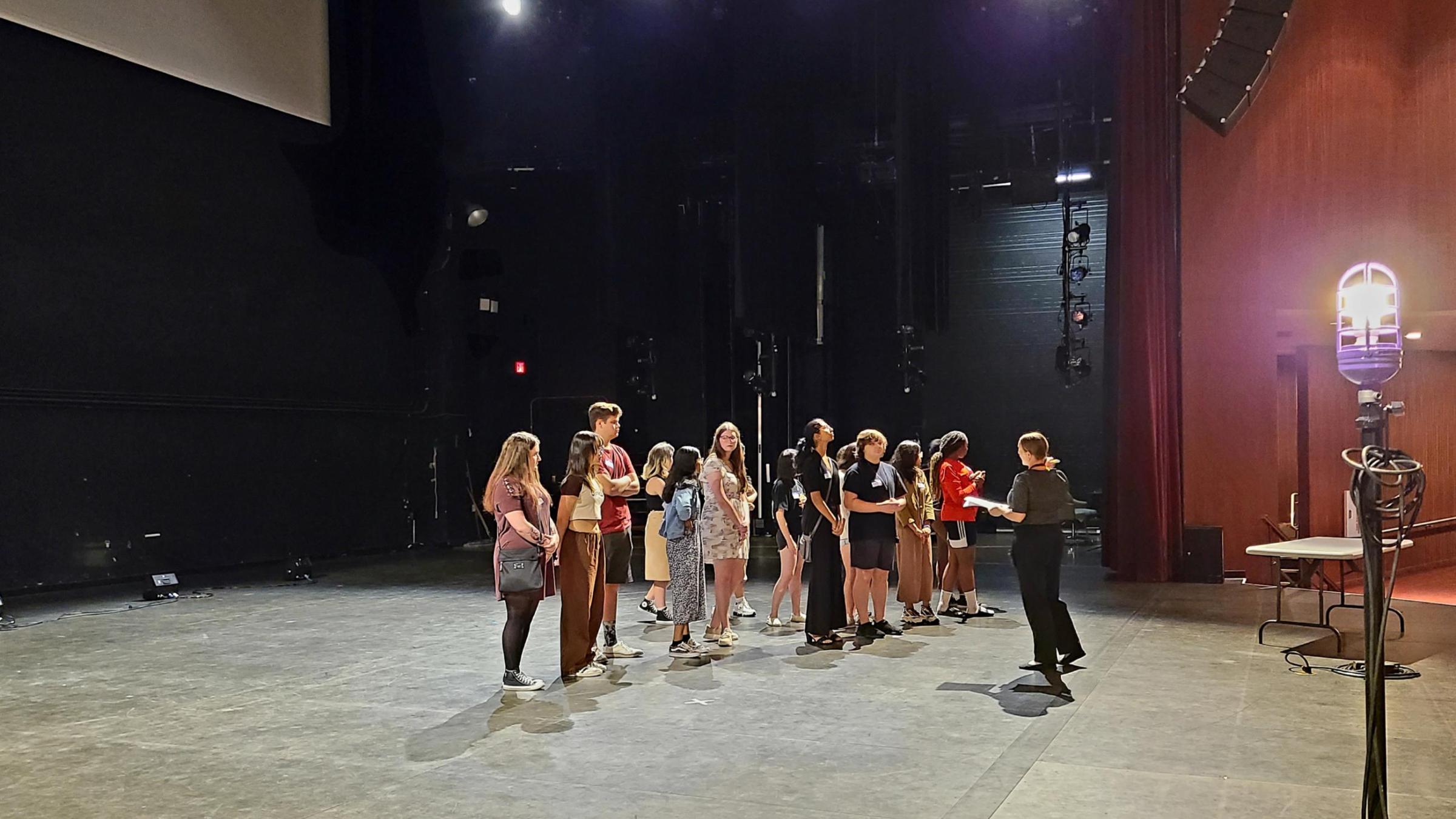 A group of students stands on a stage while a woman talks to them
