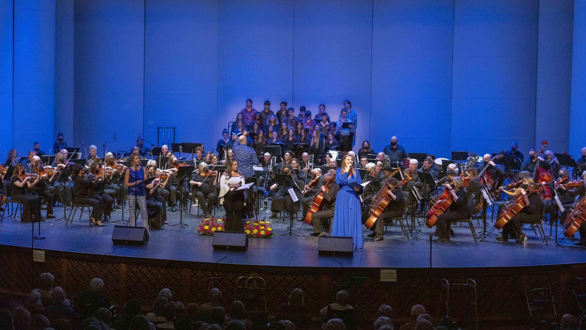 A full orchestra of musicians, conductor and two singers perform on the stage,